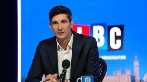 Tom Swarbrick in a navy suit jacket and white shirt, sat in front of a black microphone and with an earpiece in. In the background is a blue background with the outline of Westminster Palace visible and a red, blue and black logo that reads LBC.