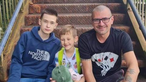 Jaroslaw Rossa and his sons Filip and Dominic - Jaroslav is wearing glasses and a black T-shirt with a design showing playing cards on it.  Dominic is sitting in the middle holding a stuffed toy and Filip is wearing jeans and a blue top. They are sitting on steps at a train station.