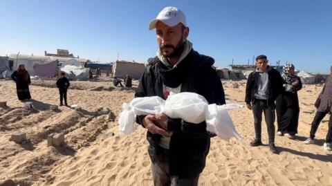 A man in a cap holds a small package wrapped in cloth containing the body of his dead newborn