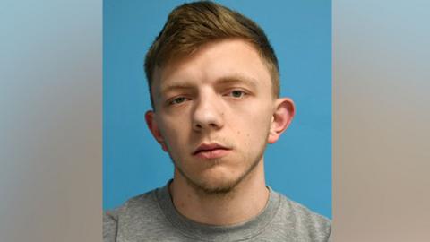 A mugshot of a man with blonde hair and a blonde beard. He wears a grey top against a blue background.
