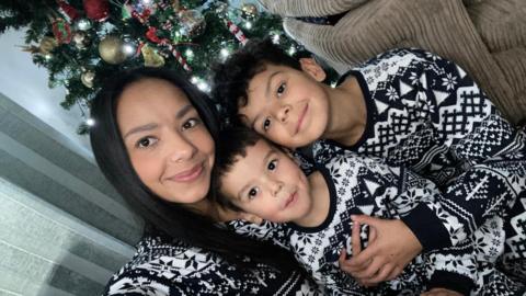 A mum takes a selfie with her two young sons in front of their Christmas tree. They are wearing matching black and white festive pyjamas.