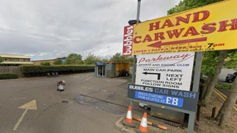 A large yellow sign for Bubbles hand car wash outside its entrance. It is on the right and beneath it is a white sign saying Parkway Sports and Social Club. On the left is a road with a faded painted arrow, beyond it a shed and beyond that a hedge above which are retail park building roofs. 