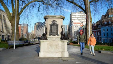 Empty stone plinth, with ornate metal detailing on the corners, and metal plaques on the flat sides, which cannot be read in the image. Lots of tall buildings in the background and two people wearing warm clothes. A large tree is on the left of the image, and it has no leaves, indicating it is winter.