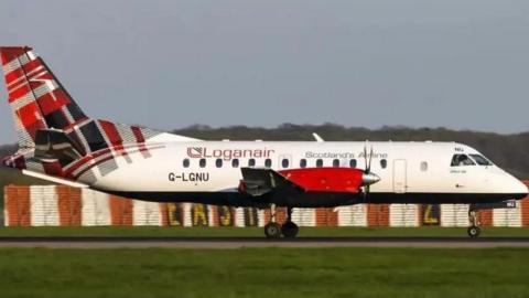 A Loganair aircraft on a runway with grassy areas either side. It is a white plane with Loganair written on the side and and a red tartan pattern on the tail.