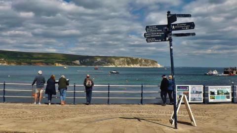 A photo of people standing against a railing looking out to sea, with a signpost in the foreground, cliffs in the background and a mix of clouds and blue sky overhead