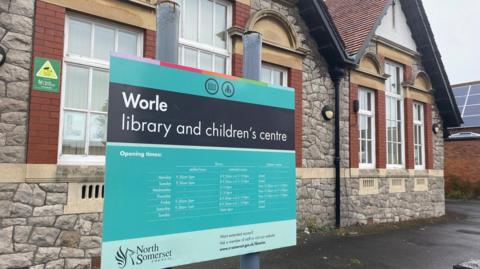A blue and black sign outside Worle Library. The building is made of bricks with tarmac around it.