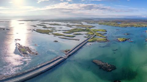 An aerial view of a causeway linking islands in the Western Isles. The causeway crosses an area of calm, shallow area of sea dotted with grassy islets.