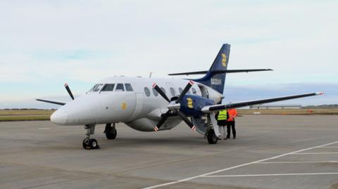 A Jetstream J32 aircraft sat on the runway. It's a grey plane with navy blue and yellow wings and black propellers with red and white tips.