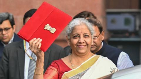 India's Finance minister Nirmala Sitharaman wearing an eggshell coloured saree with a golden border with black prints and a red blouse poses for a photograph with a red packet with India's emblem on it as she leaves the ministry of finance to present the annual budget at the parliament in New Delhi on February 1, 2025. 