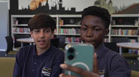 Two boys in school uniform look at a mobile phones. School shelves are in the background. 