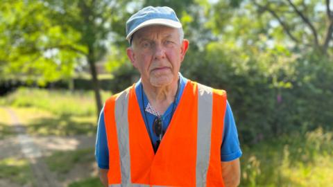 Steve Battle is wearing a blue cap, a bright blue polo shirt and an orange hi-vis jacket. He has a sad expression.