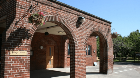 Exterior view of the chapel at the entrance to Chanterlands Crematorium. The building has a large set of wooden doors and a bricked arched covered canopy with a hanging basket of flowers attached to the front.