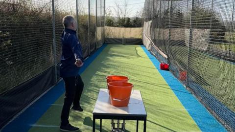 A man bowling down a green stretch of astro with two red buckets and netting in place to stop the ball from going too far.