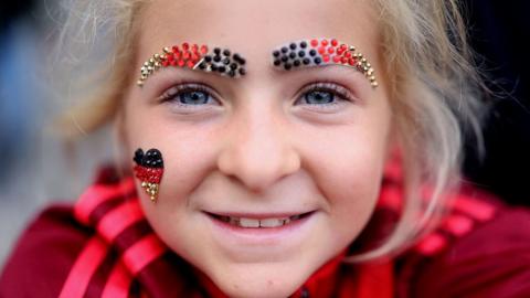 A young girl with blonde hair and blue eyes smiles to the camera in a close up shot. She is wearing sparkly eyebrows made out of beads in the colours of the German flag and a little heart on her right cheek.