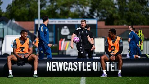 Newcastle United coach Eddie Howe (centre) carrying a ball as he looks on at the club's training ground in August. He is wearing a black club outfit. Players Malick Thiaw (left) and Jacob Ramsey are sitting down. They are wearing shorts, T-shirt and orange vests.