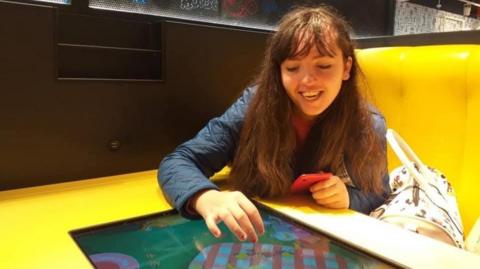 A girl with long brown hair and a blue jacket sits on a yellow restaurant booth chair at a yellow table, with a white bag next to her. She is smiling while touching a touchscreen on the table.