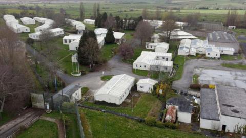 Several single-storey white buildings behind a high fence, as seen from above.