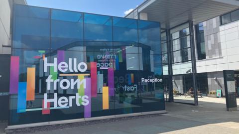 A picture of a glass reception area at the University of Hertfordshire in Hatfield. It has the words "Hello from Herts" on the outside and some colourful blocks of solid colour in red, blue and orange next to the lettering.