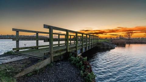 A wooden bridge crosses a small body of water and in the background there are some trees on the horizon with horizontal streaks of yellow and orange in one half of a twilight sky