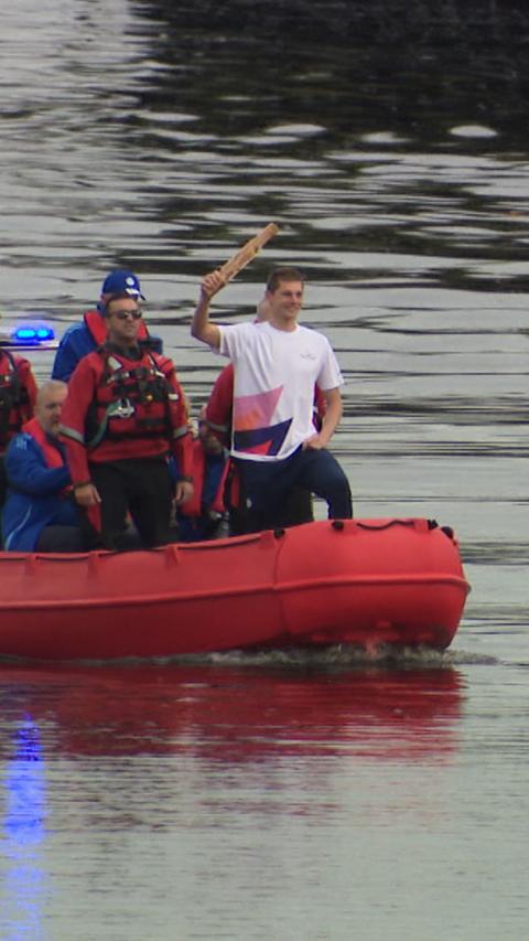 The swimmer Duncan Scott holding the King's Baton in the air. He is standing on a boat in the River Clyde