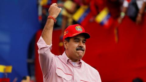Nicolás Maduro gestures during a ceremony in Caracas on 1 December, 2025.
He is wearing a red-and-white striped shirt and a re baseball cap. He has lifted one arm in the air. 