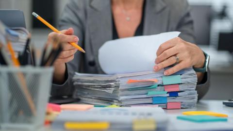 A close-up of a woman flicking through a stack of files with multi-coloured post-it notes in it. She is wearing a grey suit jacket