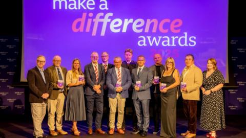 The winners from the 2025 Make a Difference Awards in Kent. Twelve people are stood in a line on a stage, in front of a projector which says: "Make a difference awards 2025"