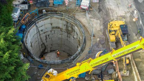 An overhead drone shot of a deep, circular concrete hole which is the tank. Two workers in orange high-vis clothes are standing at the bottom of it. Around the top is a construction site with large metal fencing, branches of a tree and a large yellow crane arm.