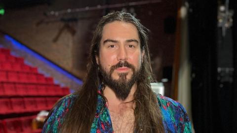 Adam has long brown hair and a beard. He is wearing a multi-coloured top.