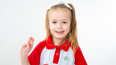 Little girl wearing a red and white Rainbows polo shirt with a Girl Guiding badge on it. She has white ribbons in her blonde hair and is holding up her right hand with three fingers extended - palm towards the camera - symbolising the Girl Guide salute.
