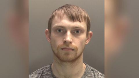 A police-issued mugshot of Kieran Williams. He has a neutral expression, fair short hair and a double bottom lip piercing. He is wearing a grey t-shirt.