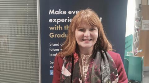 Shows a woman in red hair with a read coat and scarf and smiling