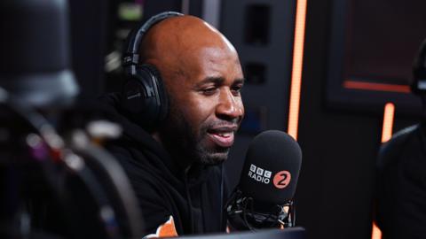 DJ Spoony speaking into a large microphone with the BBC Radio 2 logo. He is wearing headphones and smiling.