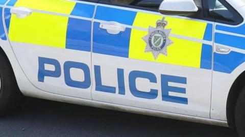 A close-up of the side of a police car. It is white with bright blue and yellow decor. The word "police" is printed in large blue letters. A Lincolnshire Police badge - star topped with a crown - sits on the door. 