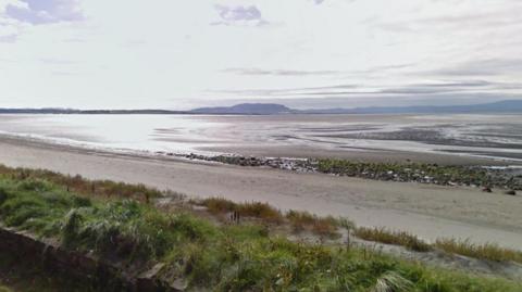 A sandy beach at low tide, with the beach going into the water in the distance. Hills are visible in the background. It is a sunny day, with sparse clouds in the sky.