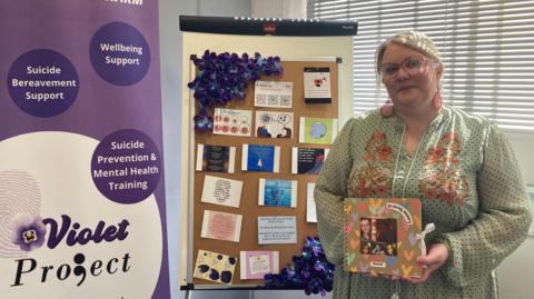 There is a purple and white upright banner with slogans about the violet project on the left, in the middle there is a cork board with some postcards on and on the right is the founder of the charity, Mel Griffin, who has blonde hair and is wearing glasses and a long light green dress. She is holding a memory book dedicated to her younger sister. 