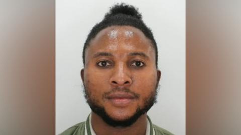 A police custody image of a man. He has short black hair, a black beard and is wearing a green jacket with white stripes around the collar.