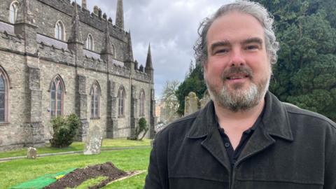 A man with curly grey hair and a grey beard stands in the foreground of a churchyard, with a recently dug grave covered in soil and green fabric visible behind them. A large stone church building and gravestones are in the background, with trees and grass under a cloudy sky.