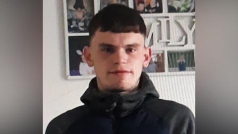 Kieran Williams who has short, brown hair and a bit of stubble on his chin. He is wearing a grey hoodie which is zipped all the way to the top. He is standing in front of a collection of photos which says 'family' in the middle. The photo is slightly pixilated, like someone has taken a photo of the picture on another screen.