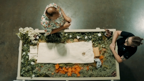 Overhead shot, showing a human composting vessel filled with flowers and organic matter, with a small white sheet laid on the top. In front of the vessel is Kristopher Hughes, wearing a colourful shirt, and opposite at the end of the vessel is one of Return Home's staff members, a women dressed in a black t-shirt with her hair tied back
