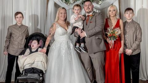 A group of six people is standing in front of a white draped backdrop decorated with a wooden arch and a floral arrangement of white and green flowers. One person is wearing a white wedding gown with lace detailing and holding hands with a child in a wheelchair covered with a cream blanket. Another person is dressed in a gray suit and holding a small child in formal attire. A person in a bright red floor-length gown is holding a bouquet of flowers. Two other individuals are wearing matching beige shirts and dark trousers. The setting appears to be indoors on a striped carpet.