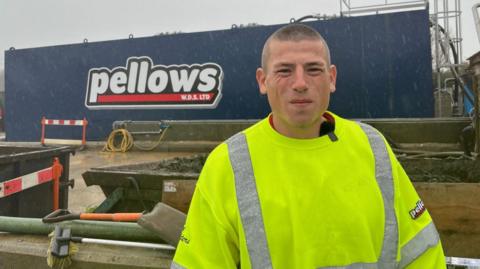 A man with a high viz  jacket stands in front of a large waste tank full of sewage. 
