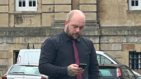 Cameron Ward, wearing a black shirt and burgundy tie, looks down at his phone outside York Crown Court