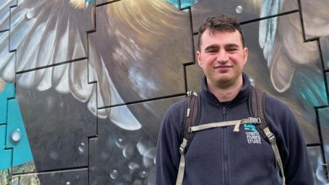 Tom Aplin, a man with dark brown hair, is wearing a navy blue fleece jacket with the Trent Valley Trust logo on the chest. He is stood next to a colourful mural of  a kingfisher.