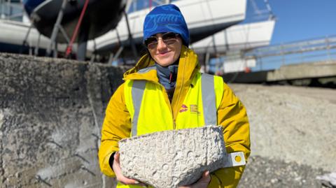 Women holding a large concrete basin. She is wearing a blue hat, sunglasses, yellow mustard coat and a yellow high vis vest with Mourne Gullion and Strangford logo. There are boats out of focus in the background and a large grey concrete wall. 