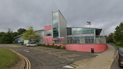 Pingles Leisure Centre, a glass-fronted building, also with an outside wall painted pink. There are parking spaces in the foreground, with one car parked. 