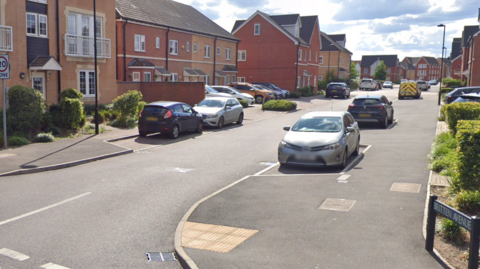The street has what look like fairly new build homes.  Parked cars are either side in bays just off of the road.  There's a few streetlights.   There's clouds in the sky but it's not overcast.