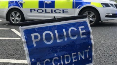 A blue police accident sign resting on the road in the foreground. In the background there is a parked police car with blue and yellow hi-vis decals.
