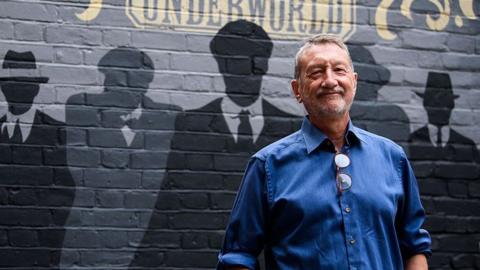 Steven Knight, creator of Peaky Blinders stands with the background of painted silhouettes of his most famous creations behind him at the opening of Peaky Blinders underworld. He is wearing a blue shirt and is smiling