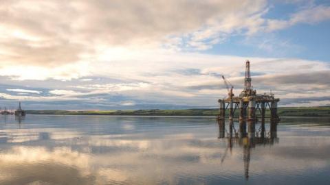 A picture of an oil rig in an estuary with a thin strip of land in the background. Thee are many clouds in the sky which are reflected in the water 